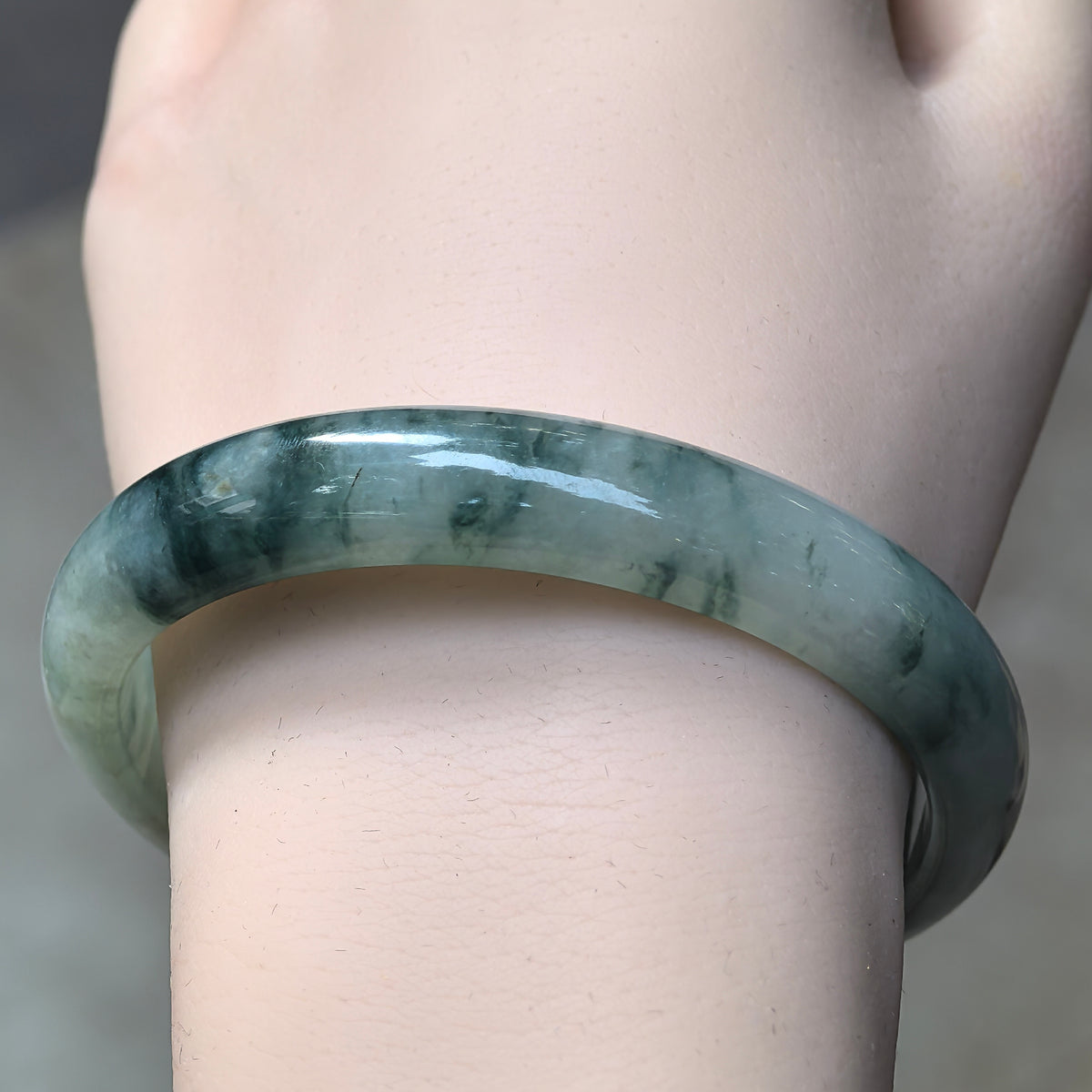 Front wrist view of a fine glutinous jadeite bangle showing scattered dark green trails and clusters within the lighter green stone.