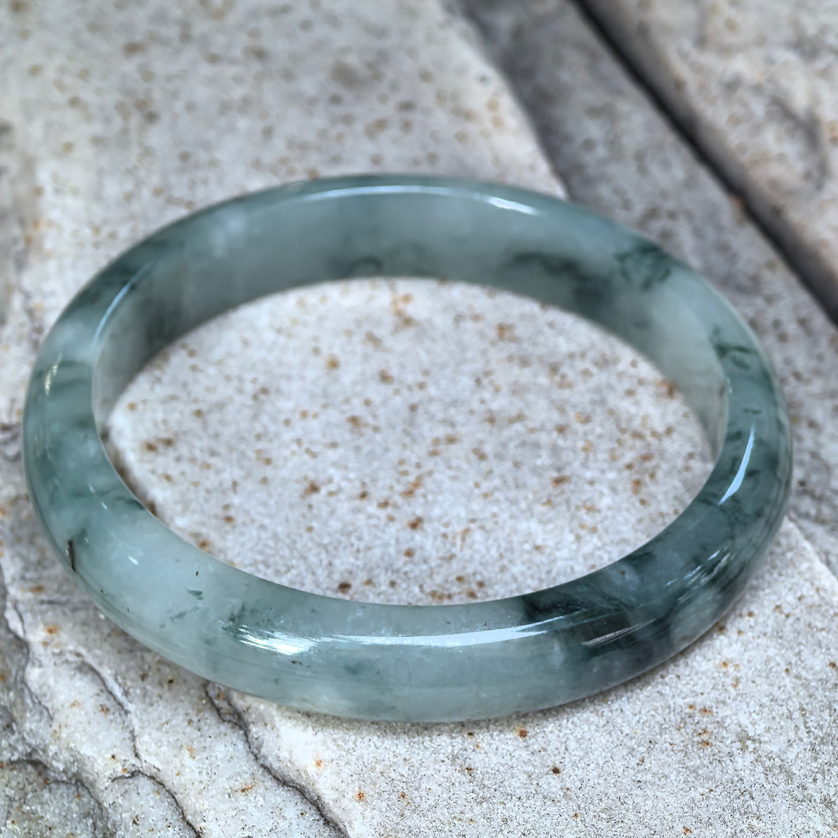 Green jadeite D-shape bangle resting at an angle on pale stone, showing its flat inner surface and a concentrated dark green section near the front.