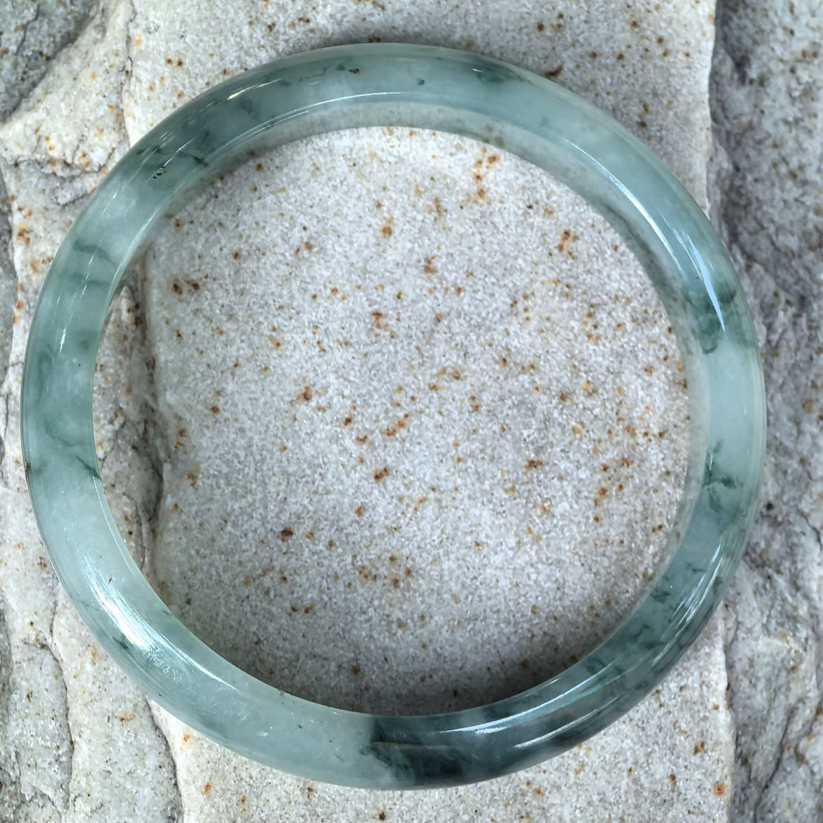 Top view of a light green jadeite bangle on textured stone, with fine dark green floating patterns distributed around the circular band.