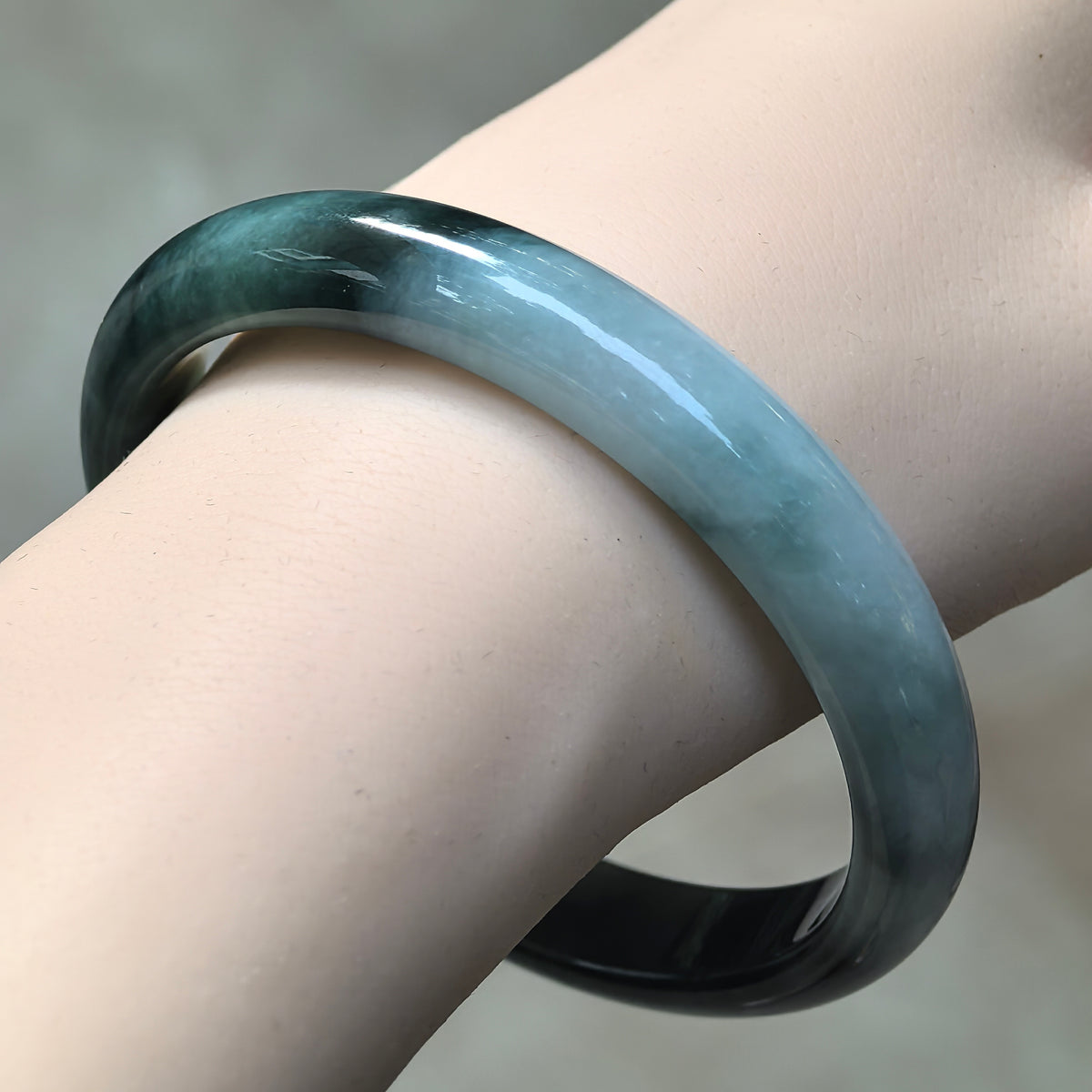 Angled wrist view of a jadeite D-shape bangle with deep green floating patterns on a light green base and a polished rounded exterior.