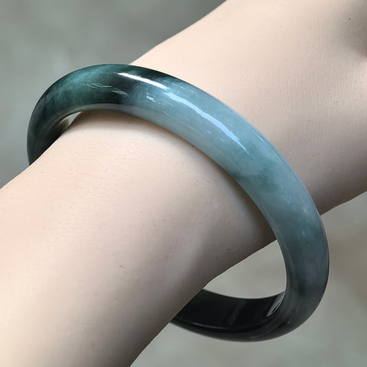 Angled wrist view of a jadeite D-shape bangle with deep green floating patterns on a light green base and a polished rounded exterior.