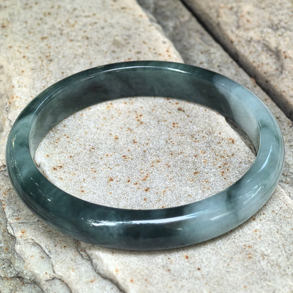 Green jadeite D-shape bangle resting at an angle on pale stone, showing its flat inner surface and contrasting light and dark green patterns.