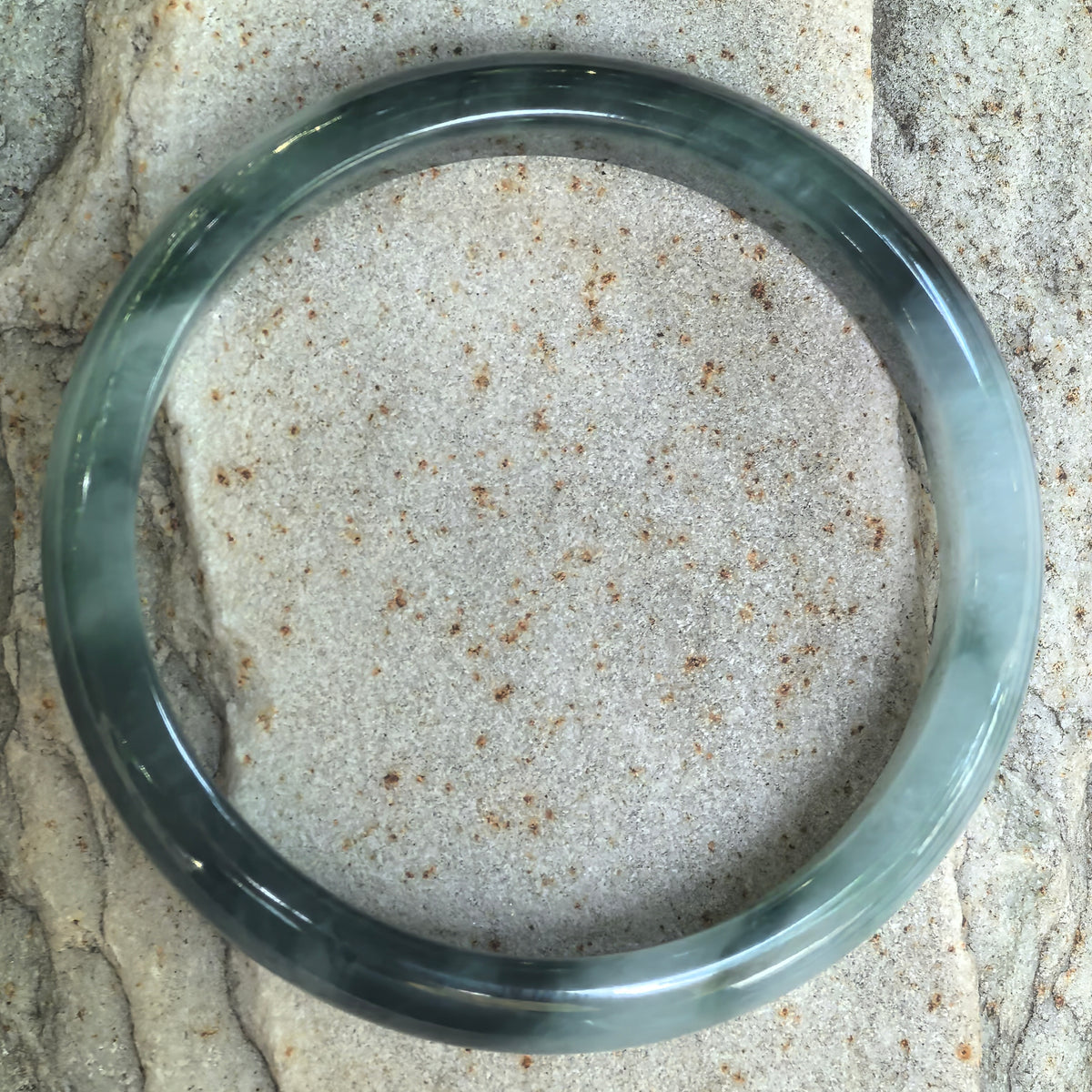 Top view of a jadeite bangle on textured stone, showing deep green floating patterns around its circular light green base.