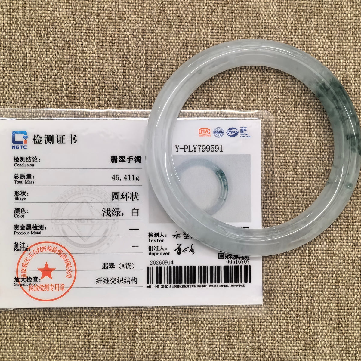 White and green jadeite round-bar bangle displayed with its matching NGTC certificate on tan woven fabric.