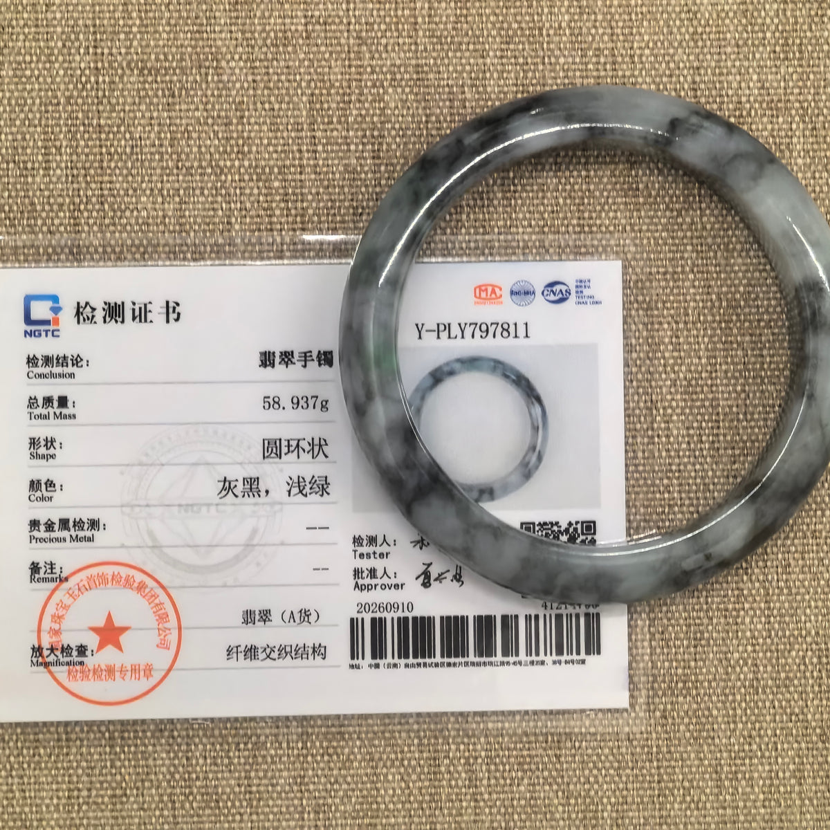 Gray and black jadeite bangle displayed with its matching NGTC certificate on a woven background.