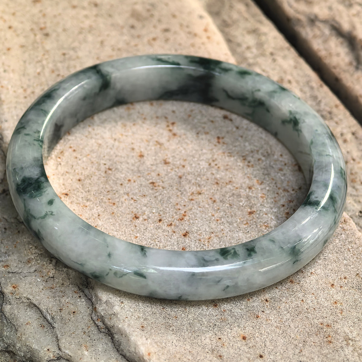 Green floating flower jadeite bangle resting at an angle on stone, showing its flatter inner face and patterns around the band.