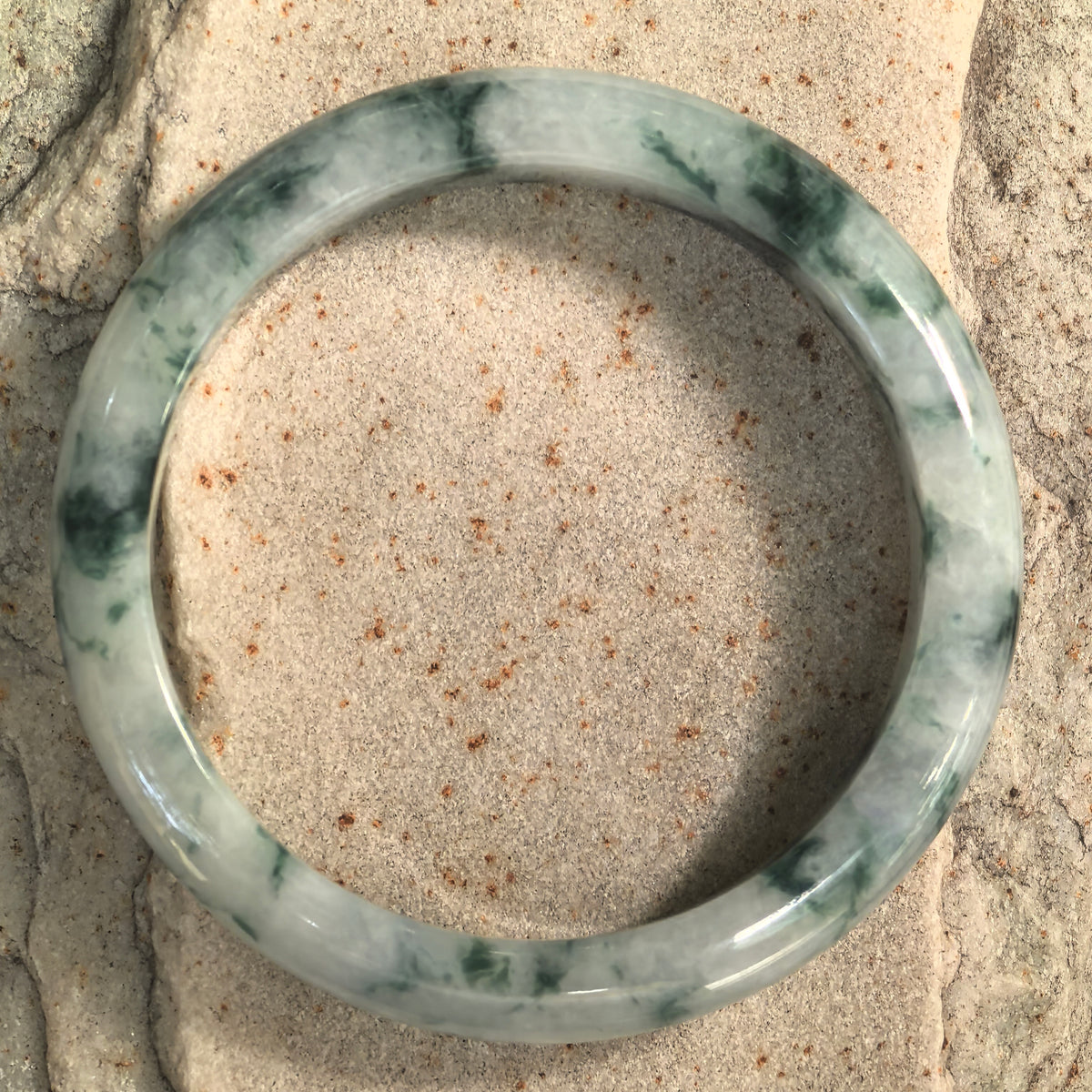 Top view of a D-shape jadeite bangle on stone, showing its circular outline and varied green markings across a milky base.