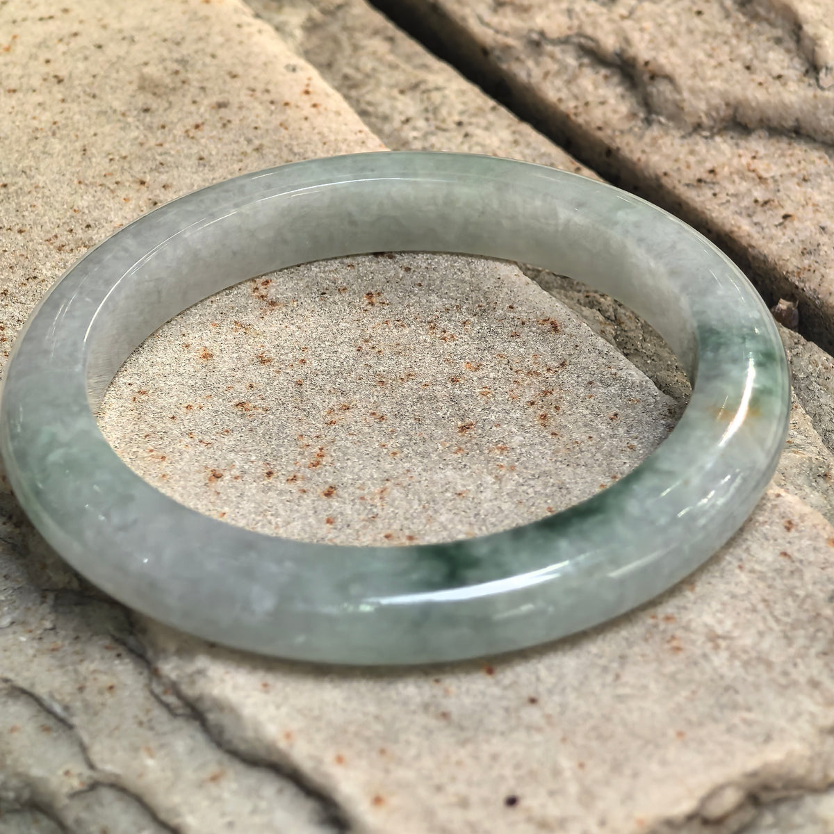 Light green jadeite bangle resting at an angle on stone, showing its flatter inner face and darker green floating patterns along the front.
