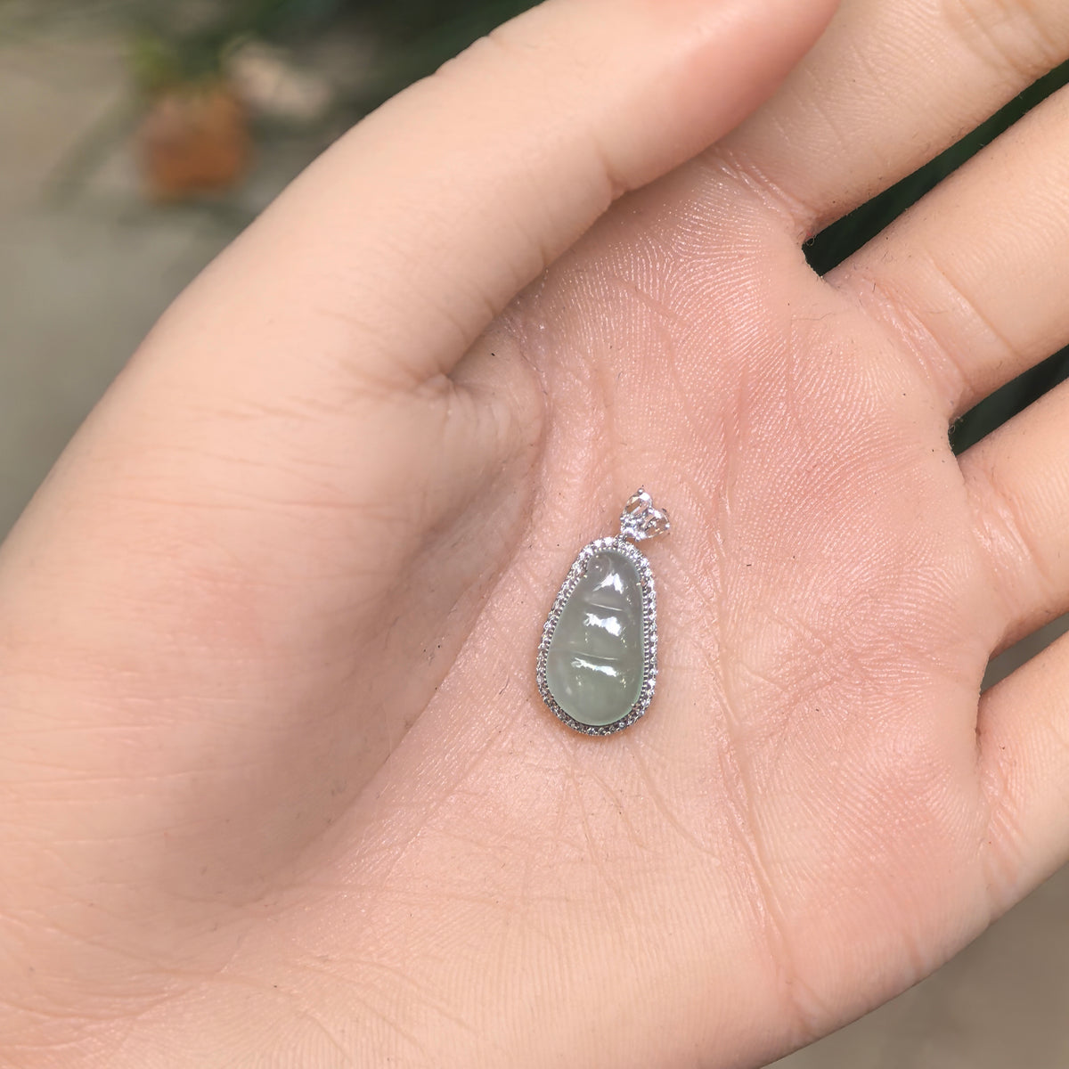Light green jadeite bean pod pendant displayed on an open palm, showing its compact proportions and 18K gold and diamond setting.