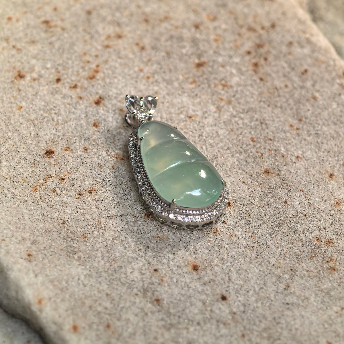 Angled view of a green jadeite bean pod pendant on stone, highlighting the rounded carving, diamond border and openwork sides of the setting.