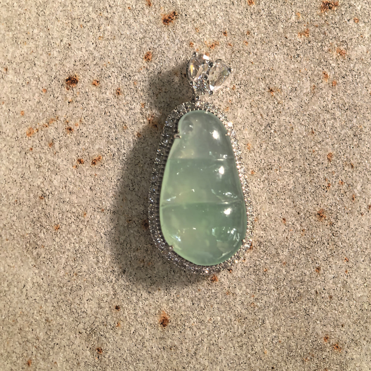 Front view of an icy light green jadeite bean pod pendant resting on stone, showing the carved segments, curled tip and paired teardrop-shaped bail details.