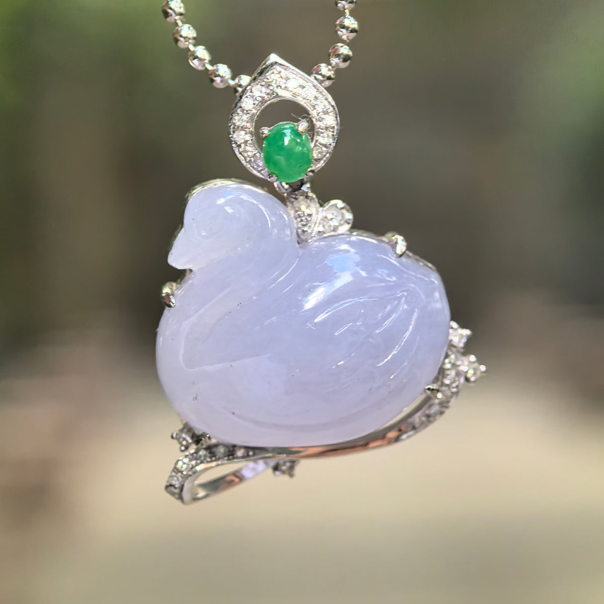 Lavender jadeite swan pendant suspended from a beaded chain, showing carved feathers, a curved diamond-accented mount and an oval green stone in the bail.