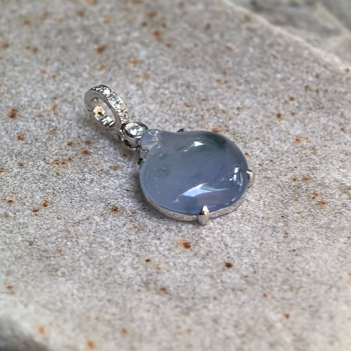 Angled view of a lavender jadeite peach pendant on stone, highlighting the rounded carving, prong setting and diamond accents above.