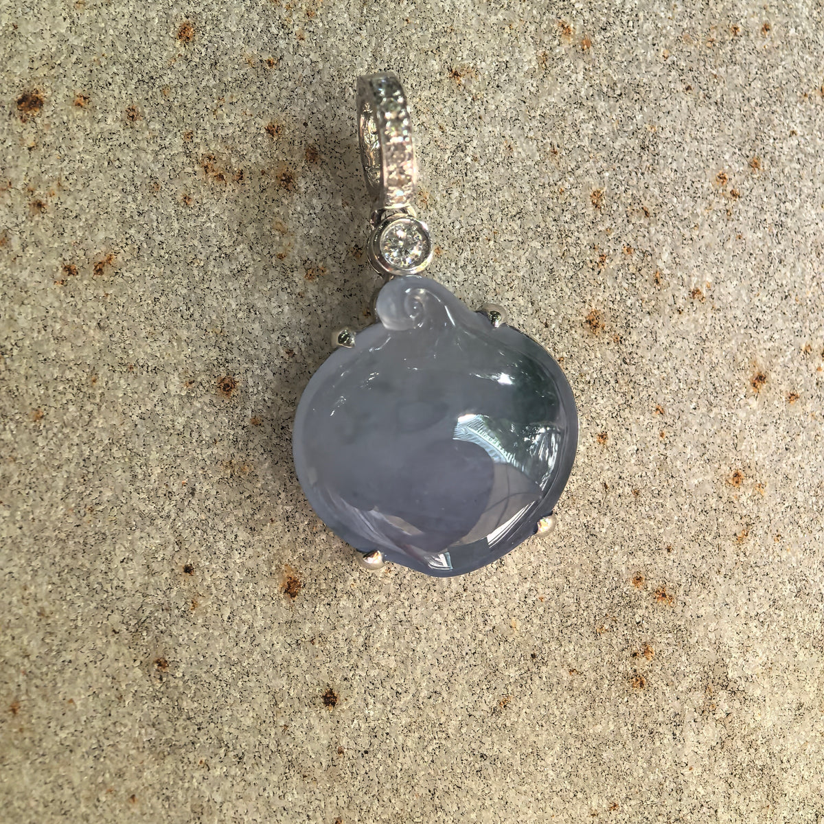 Close front view of a lavender jadeite peach pendant resting on stone, showing the localized green accent, polished surface and curled carving detail.