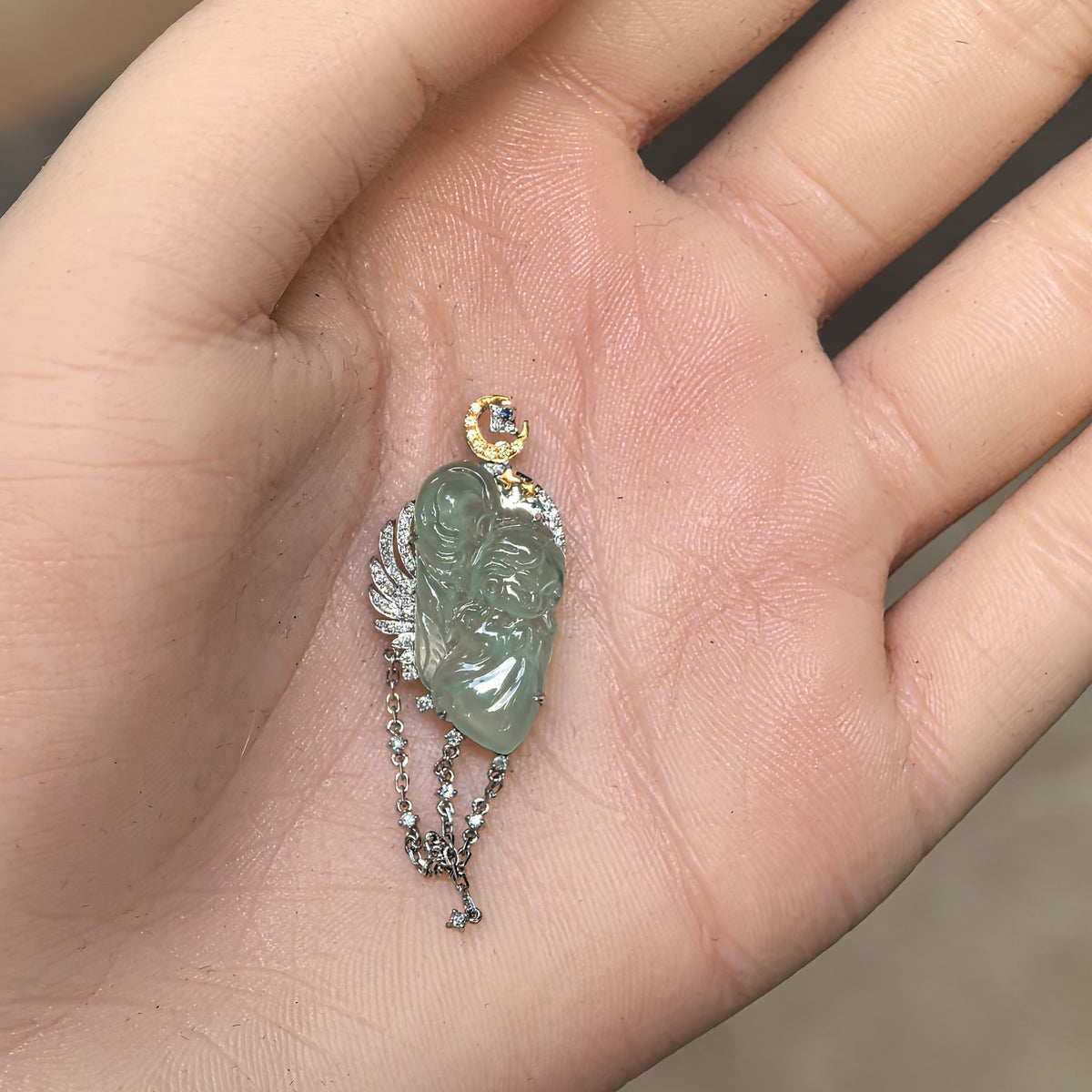 Green jadeite guardian angel pendant displayed on an open palm, showing the carved face, 18K gold setting and hanging chain details.