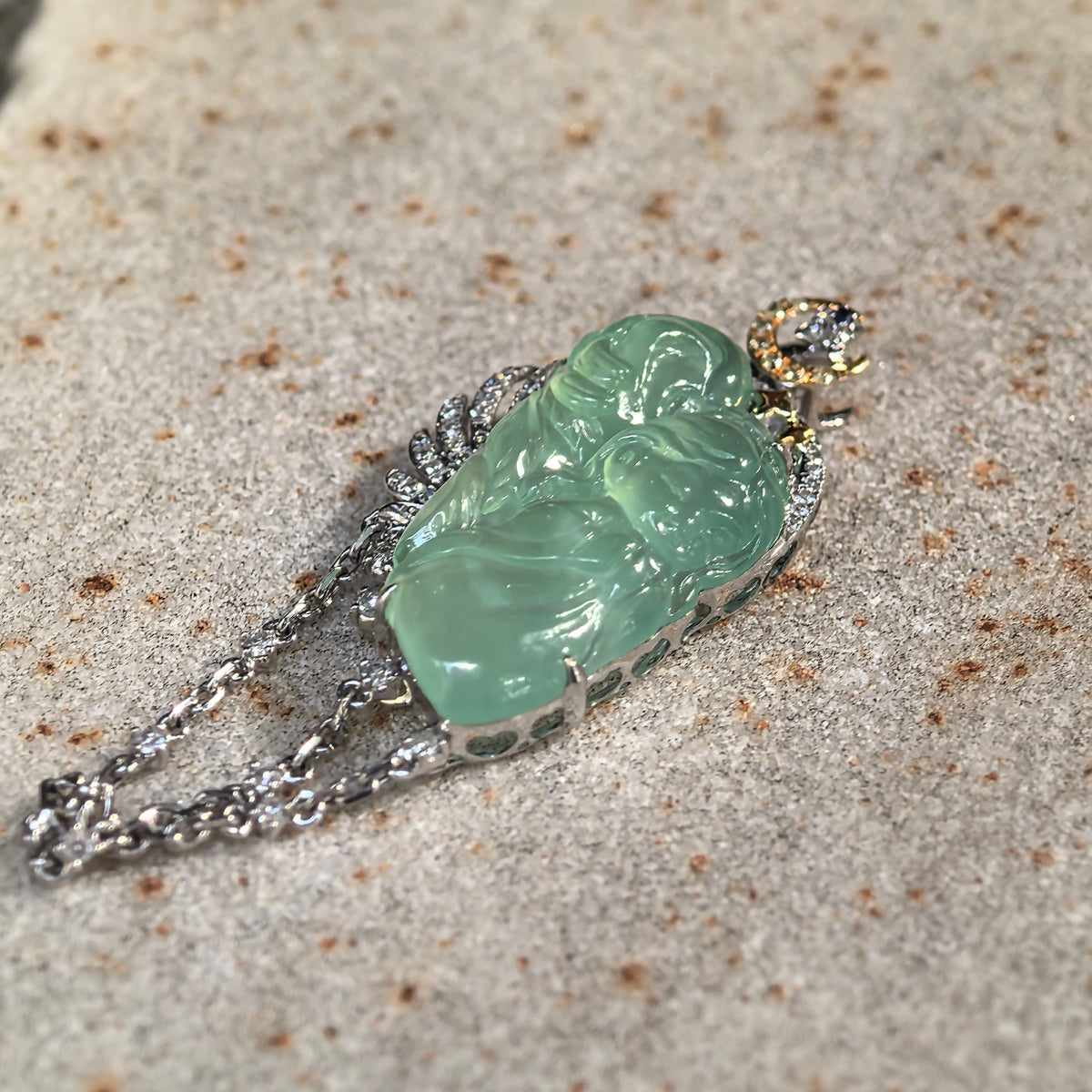 Angled view of an icy green jadeite guardian angel pendant on stone, highlighting the raised carving and openwork sides of the setting.