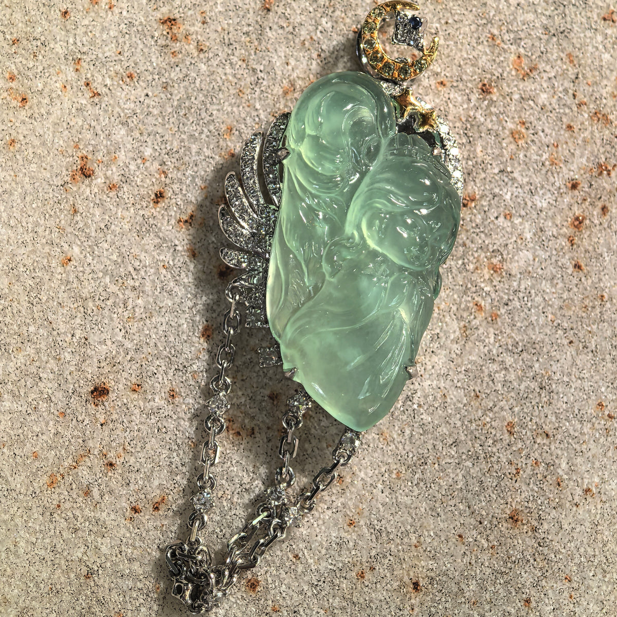 Close front view of a green jadeite guardian angel pendant on stone, showing the diamond-set wing, crescent moon, star accents and draped chains.