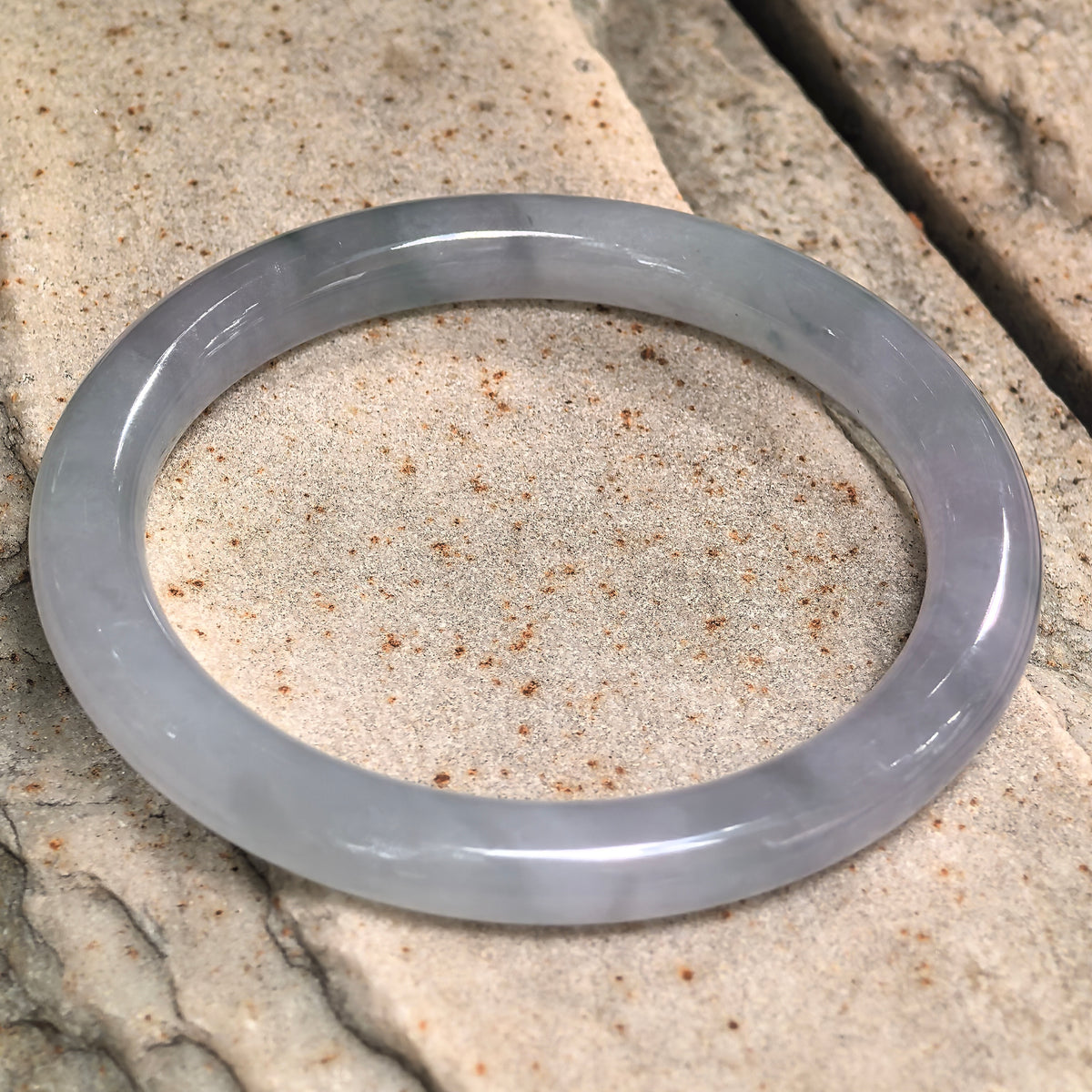 Pale lavender round-bar jadeite bangle resting at an angle on pale stone, showing its rounded inner and outer surfaces.