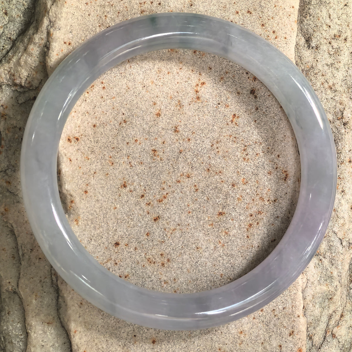 Top view of a pale lavender jadeite bangle on pale stone, showing its circular outline, subtle color variations, and polished surface.