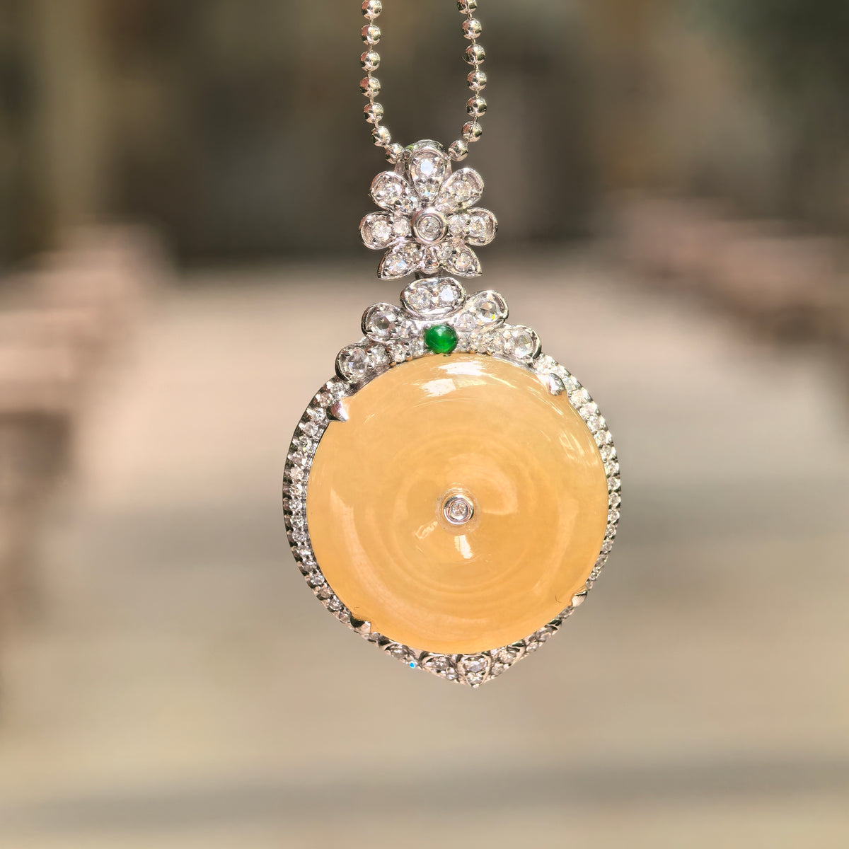 Front view of a bright yellow jadeite Peace Pendant suspended from a beaded chain, showing its diamond border, floral bail, and small green accent.