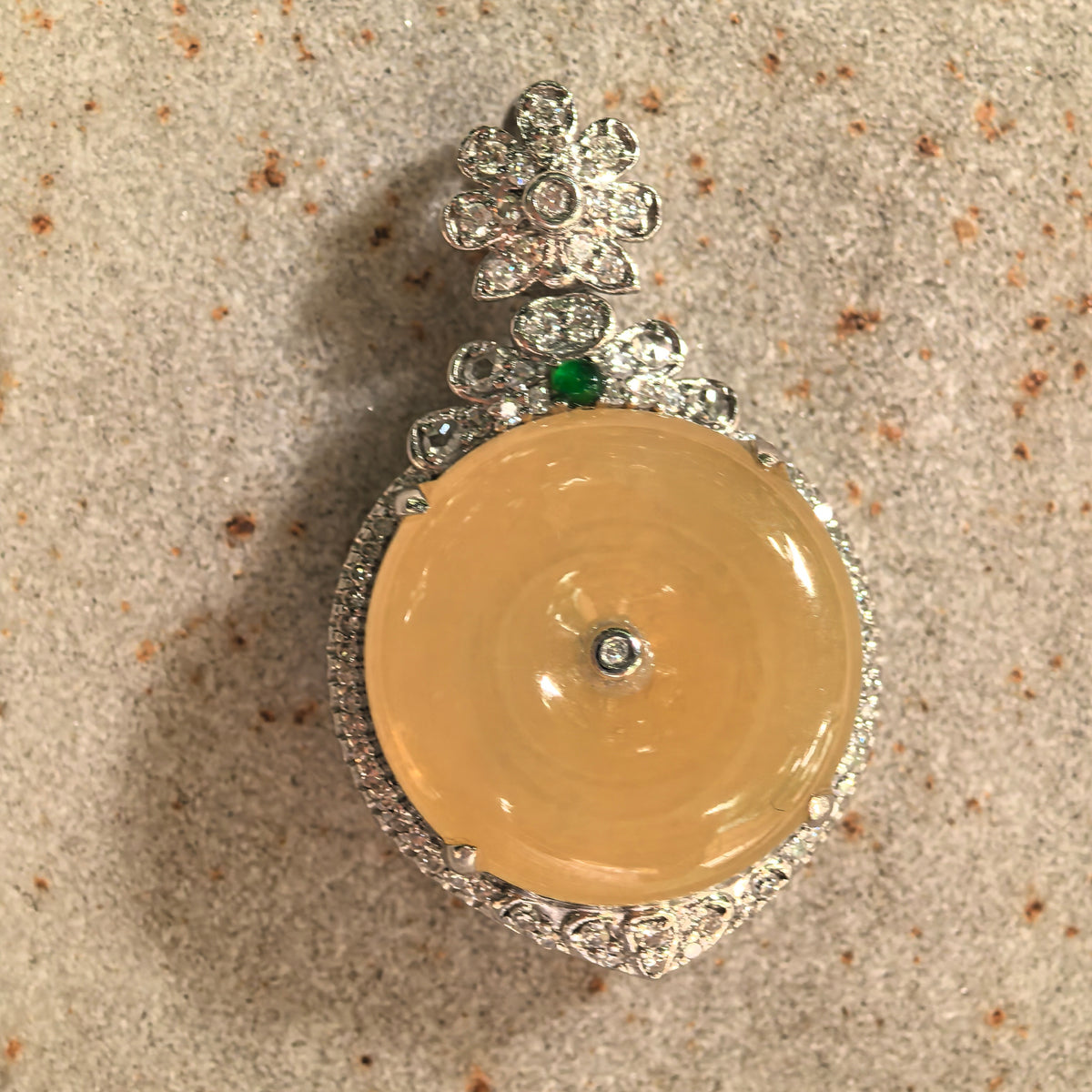 Front view of an icy yellow jadeite Peace Pendant resting on stone, with a central diamond detail and a floral bail above the green accent.