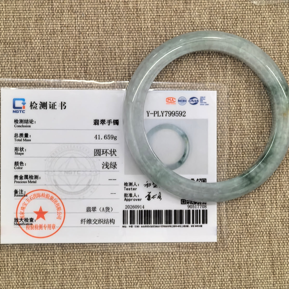 Light green floating flower jadeite bangle displayed with its matching NGTC certificate on a woven background.