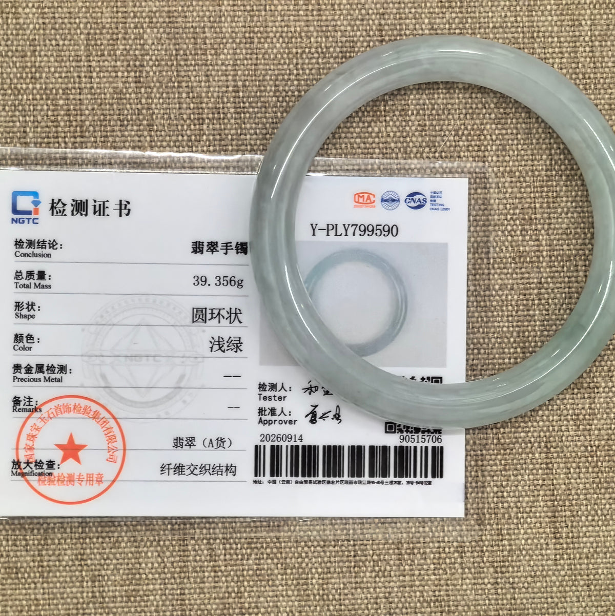 Light green round-bar jadeite bangle displayed with its matching NGTC certificate on a tan woven background.