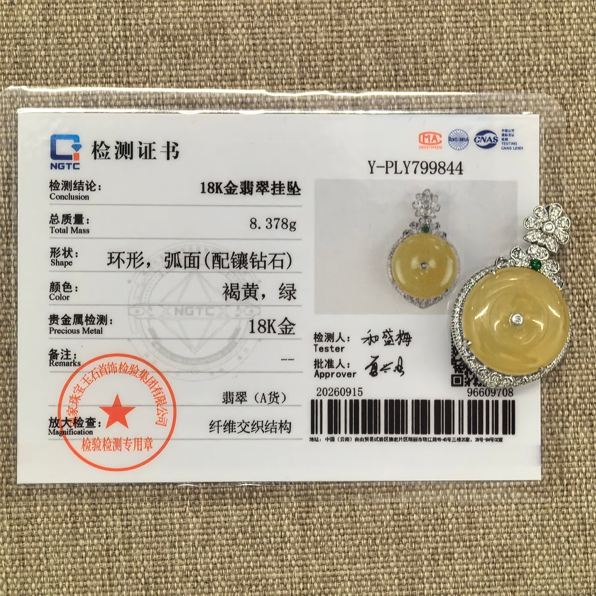 Yellow jadeite Peace Pendant in an 18K gold and diamond setting displayed beside its matching NGTC certificate on a woven background.