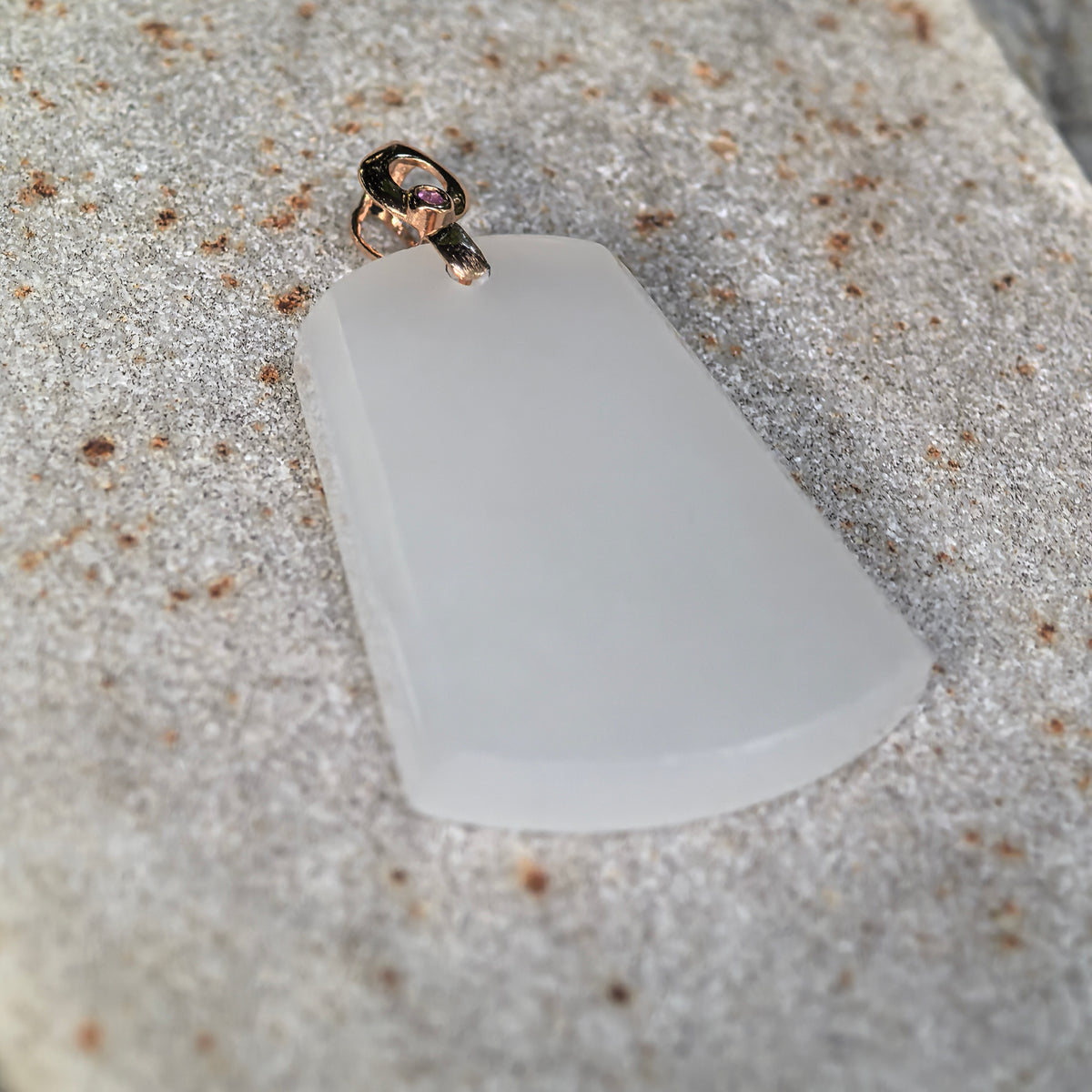 Angled view of a white Hetian jade plain plaque pendant on stone, showing the beveled edges and pink accent on the gold bail.
