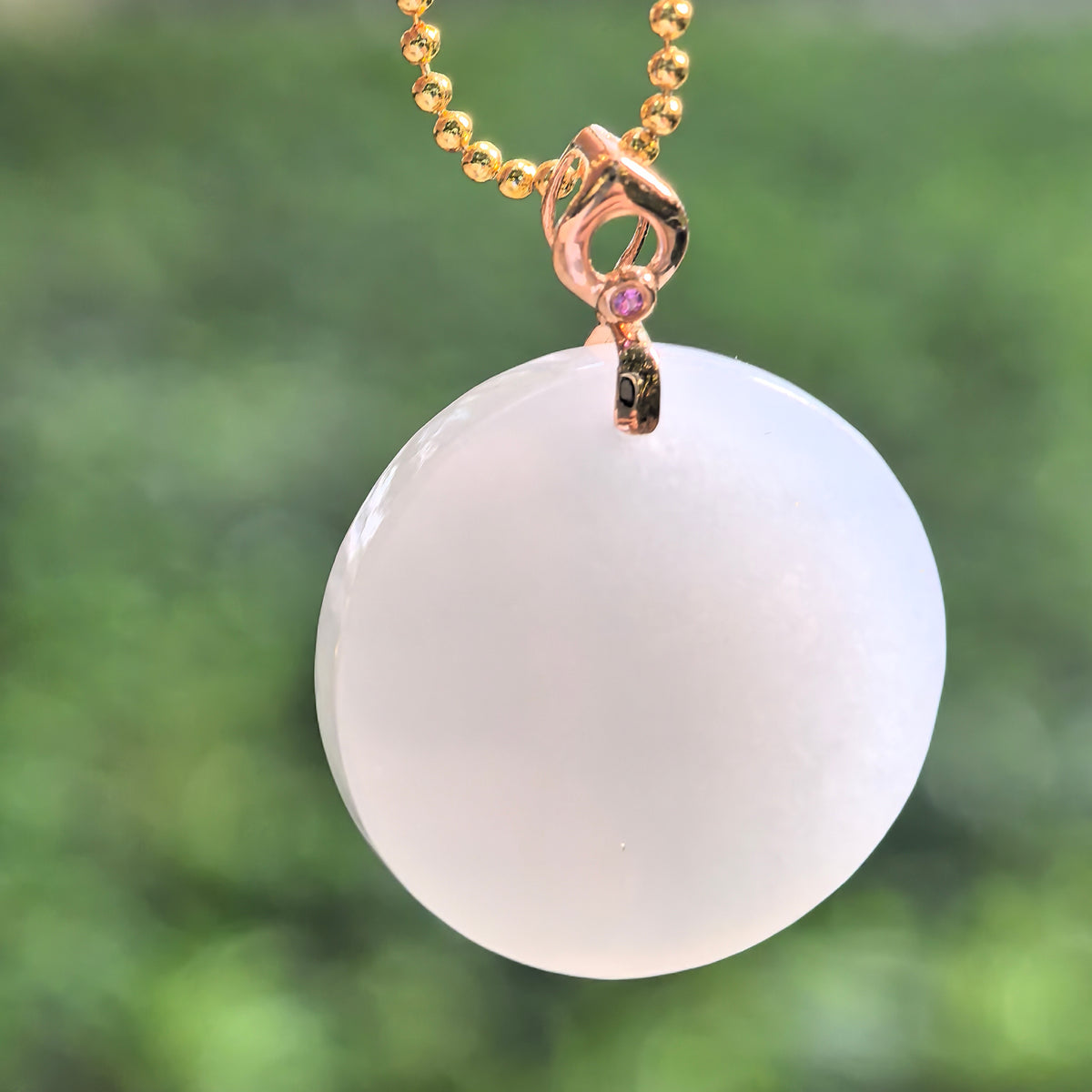 White Hetian jade round plain plaque pendant hanging from a display chain, with an 18K gold bail and a small pink accent stone.