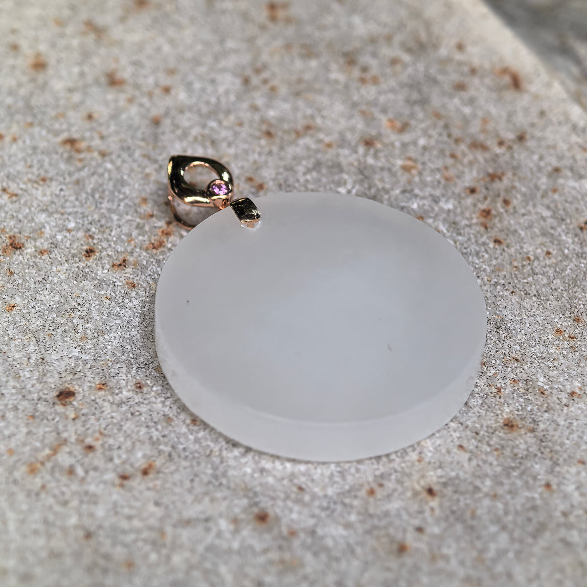 Angled view of a white nephrite round plain plaque pendant resting on stone, showing its smooth face, rounded edge and gold bail.