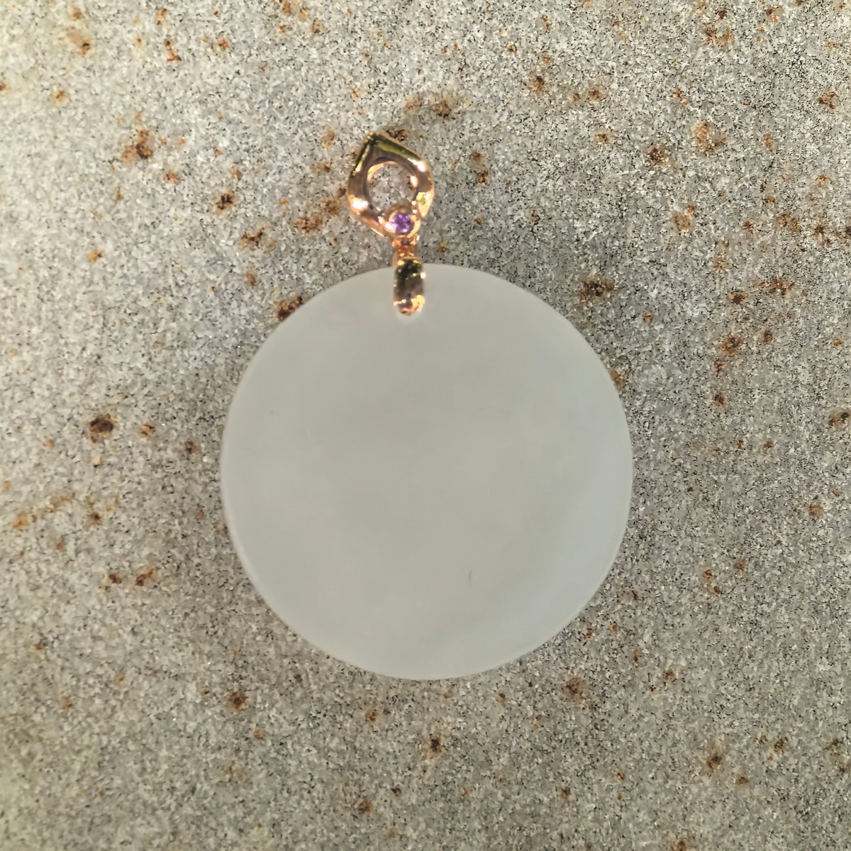 Front view of a white nephrite round plain plaque pendant on stone, with an uncarved face and a small pink accent on the gold bail.