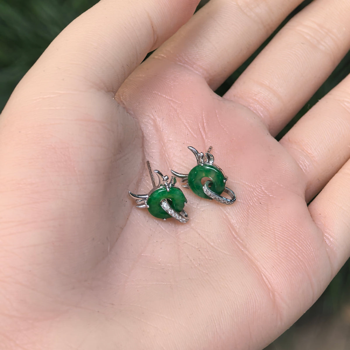 Pair of green jadeite little devil stud earrings resting on a palm, showing their size, stud posts and diamond-set loops.