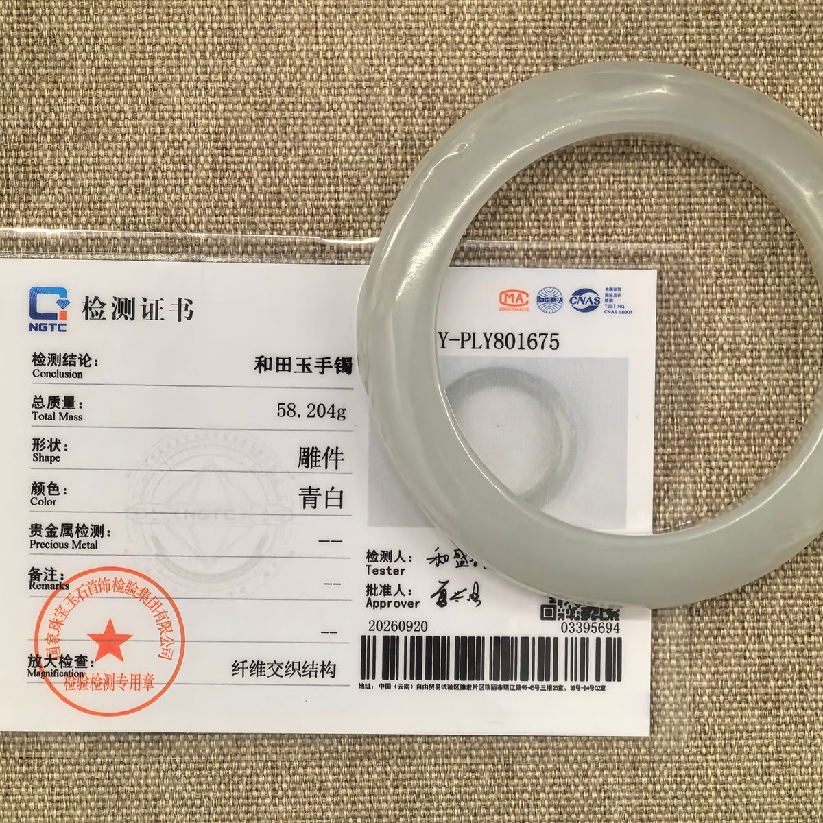 White Hetian jade lotus-carved bangle displayed with its matching NGTC certificate on a tan woven background.