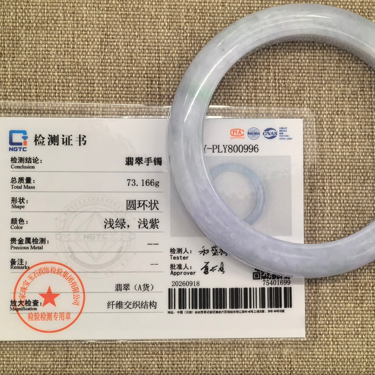 Pale lavender jadeite bangle displayed with its matching NGTC certificate on a tan woven background.