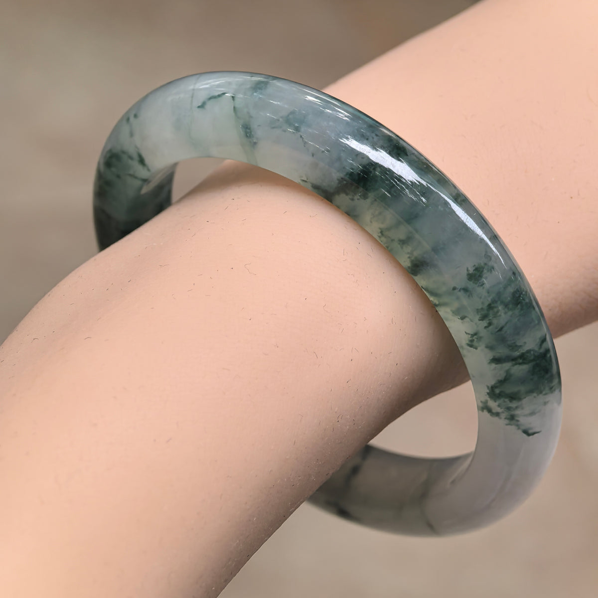 Icy glutinous jadeite D-shape bangle worn on the wrist, showing dark ink-green floating patterns across a pale gray-green base.