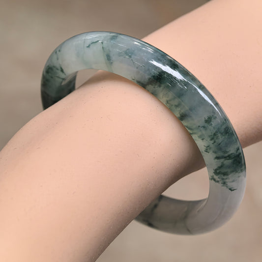 Icy glutinous jadeite D-shape bangle worn on the wrist, showing dark ink-green floating patterns across a pale gray-green base.