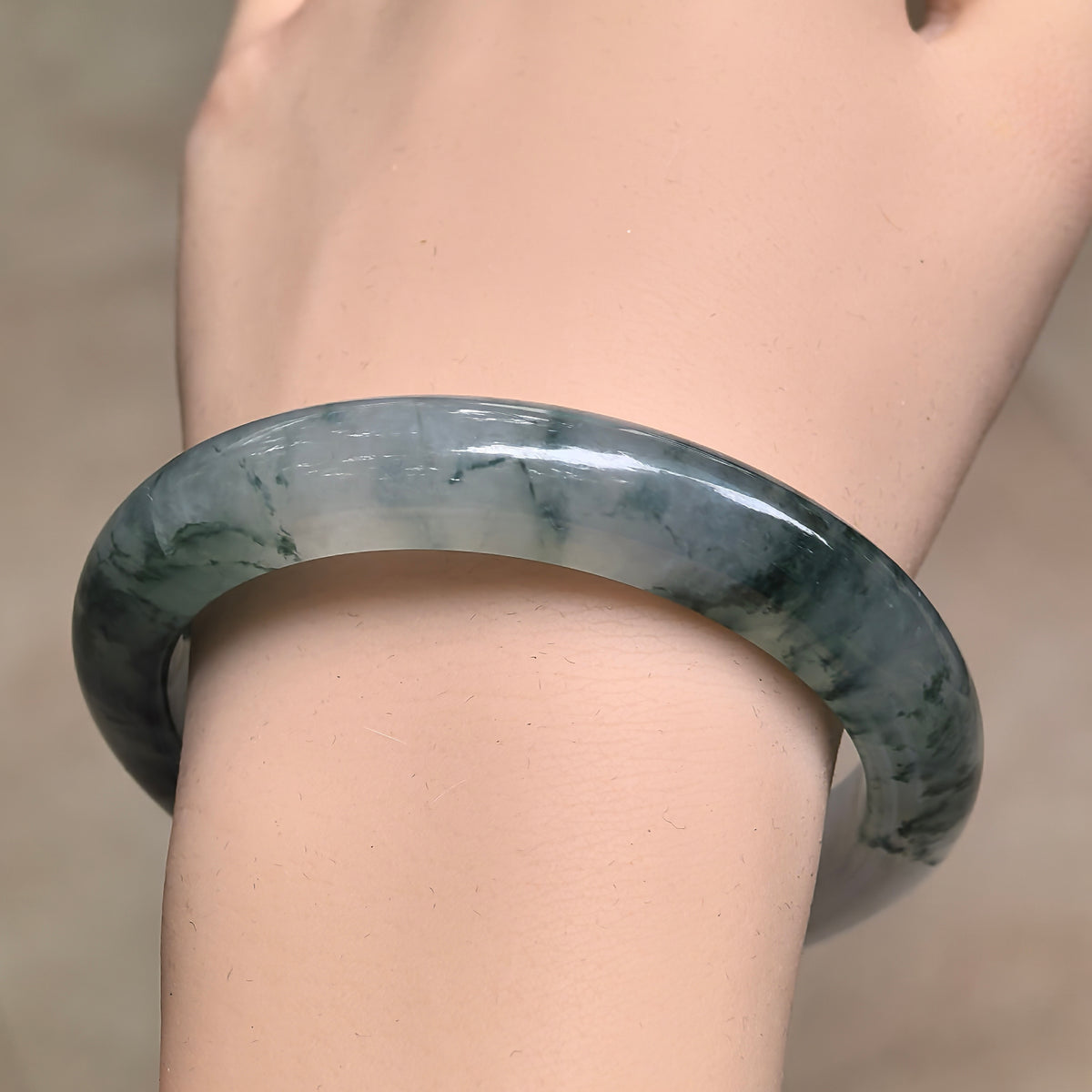 Angled wrist view of a jadeite bangle with deep green floating patterns, translucent pale sections, and a polished rounded outer surface.