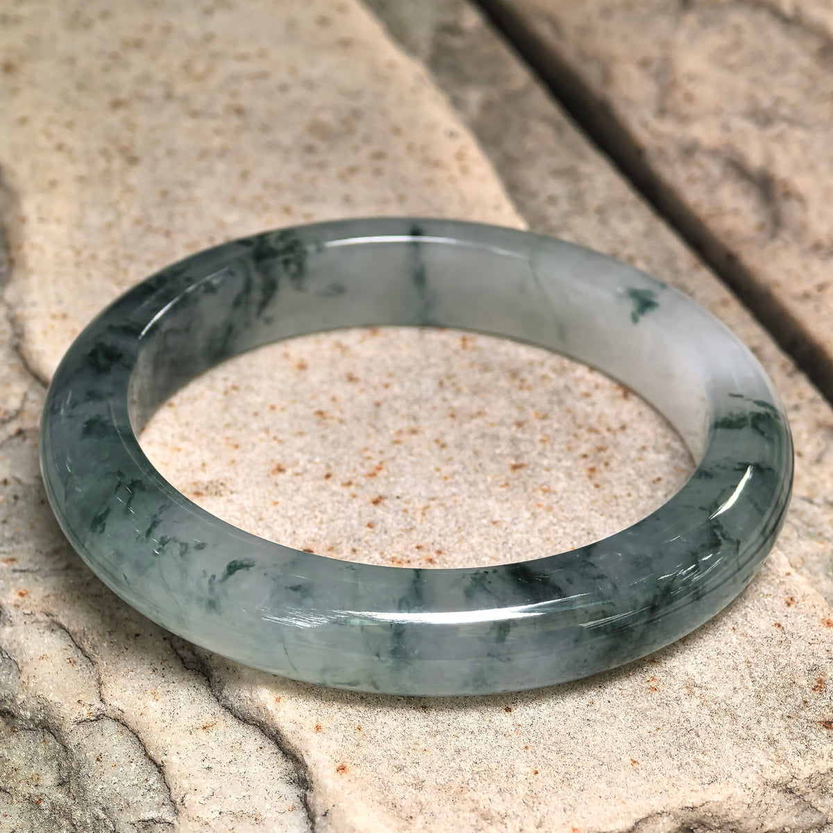 Jadeite D-shape bangle resting on light stone, showing its flatter inner face and dark green patterns through the pale gray-green base.
