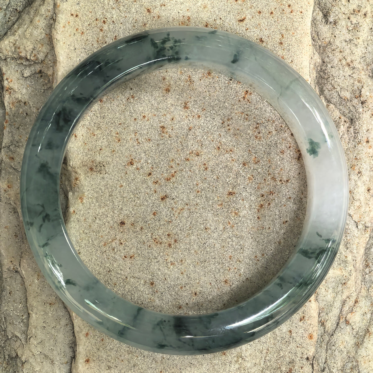 Overhead view of a jadeite bangle on stone, showing dark ink-green floating patterns and paler sections around the full circle.
