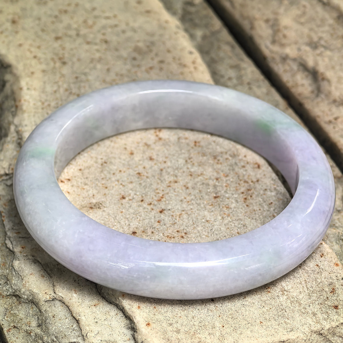 Pale lavender jadeite D-shape bangle resting at an angle on stone, with subtle light green accents and a polished surface.