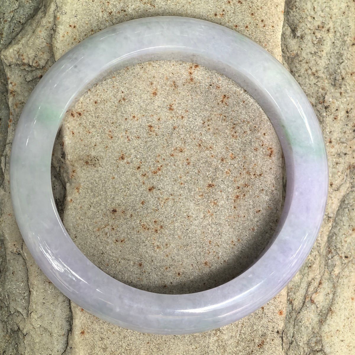 Overhead view of a jadeite bangle on stone, showing its full circular outline and natural variation between pale lavender and light green sections.