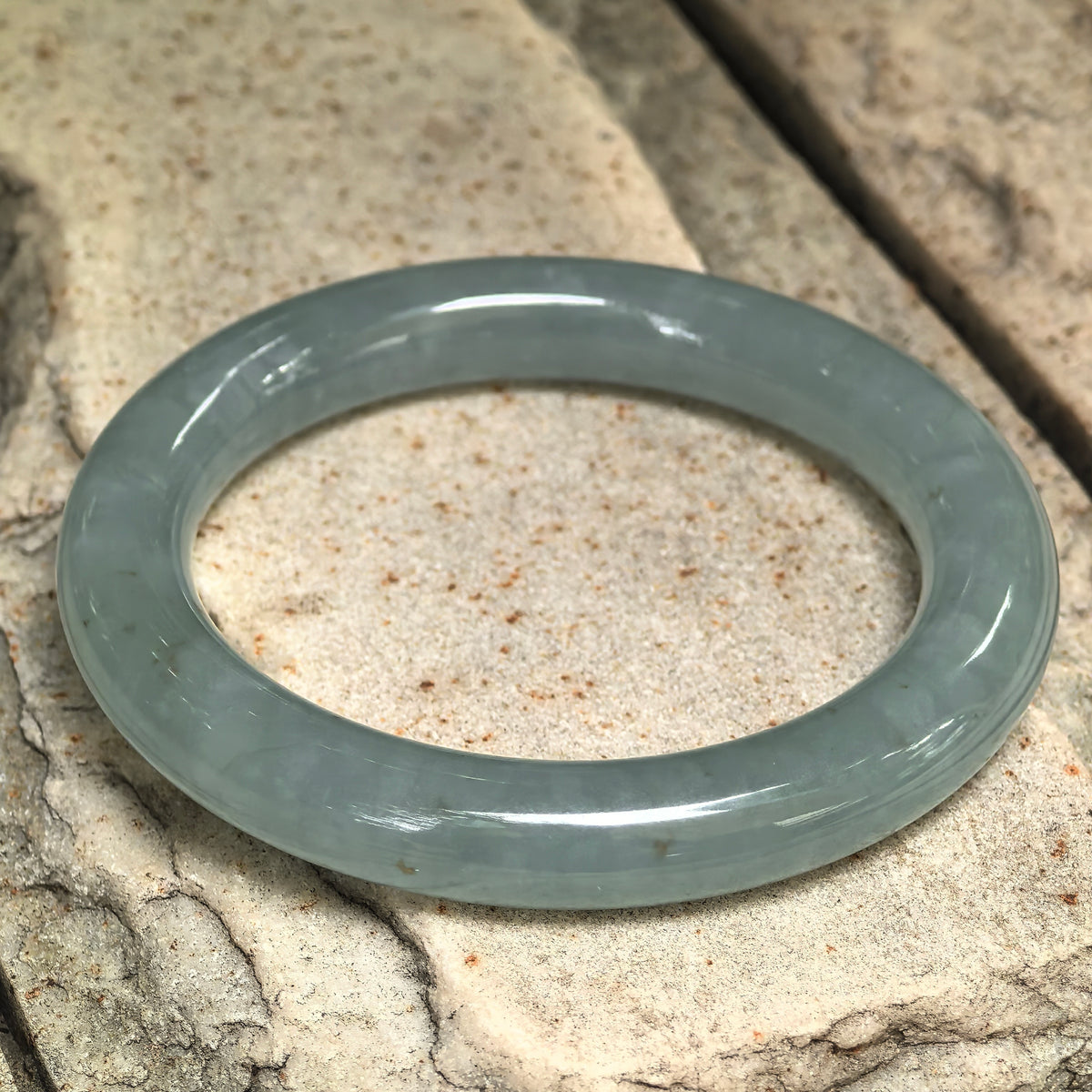 Blue-green jadeite round-bar bangle resting at an angle on stone, with small natural golden-brown flecks visible along the polished band.
