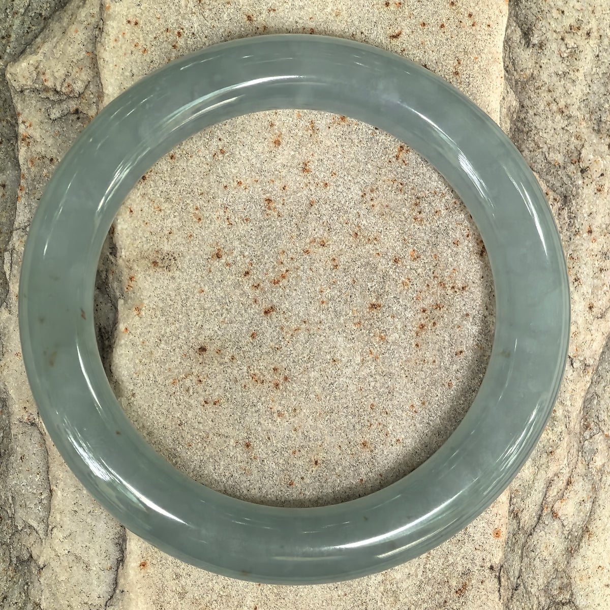 Overhead view of a blue-green jadeite bangle on stone, showing its circular outline, rounded band, and sparse golden-brown flecks.