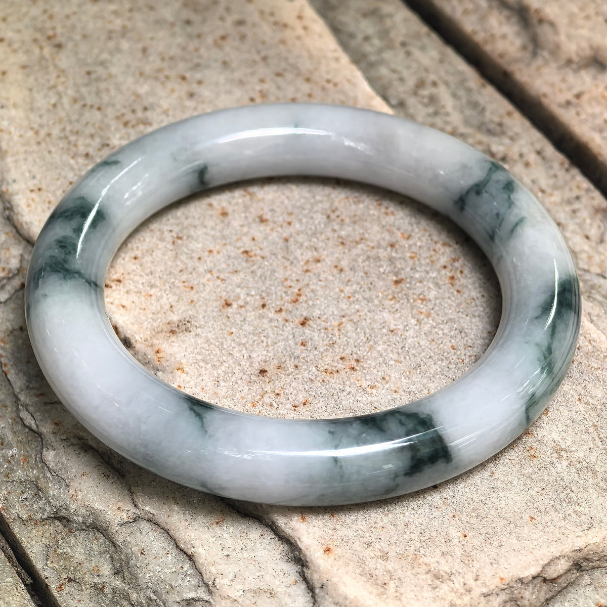 White jadeite bangle resting at an angle on pale stone, showing dark green floating patterns around the band.
