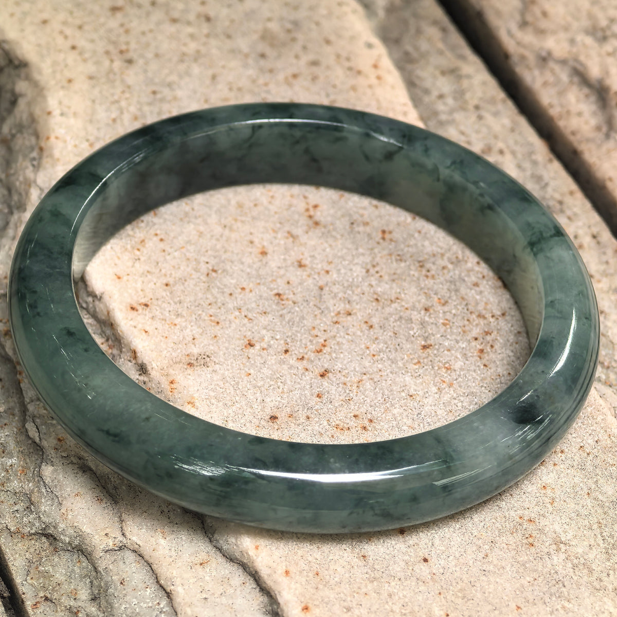 Angled view of a D-shape jadeite bangle resting on pale stone, showing its flat inner profile and varied green patterns.