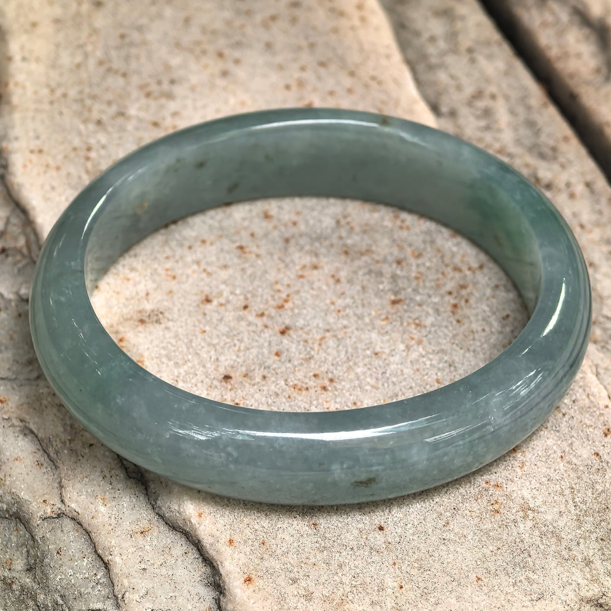 Angled view of a blue-green oval jadeite bangle resting on pale stone, showing the inner surface and rounded outer face.