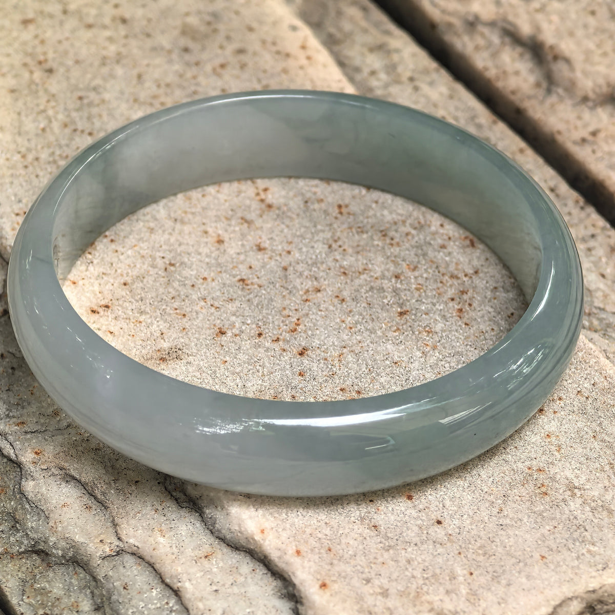 Angled view of a pale green jadeite bangle resting on stone, showing its flatter inner face and gently rounded exterior.