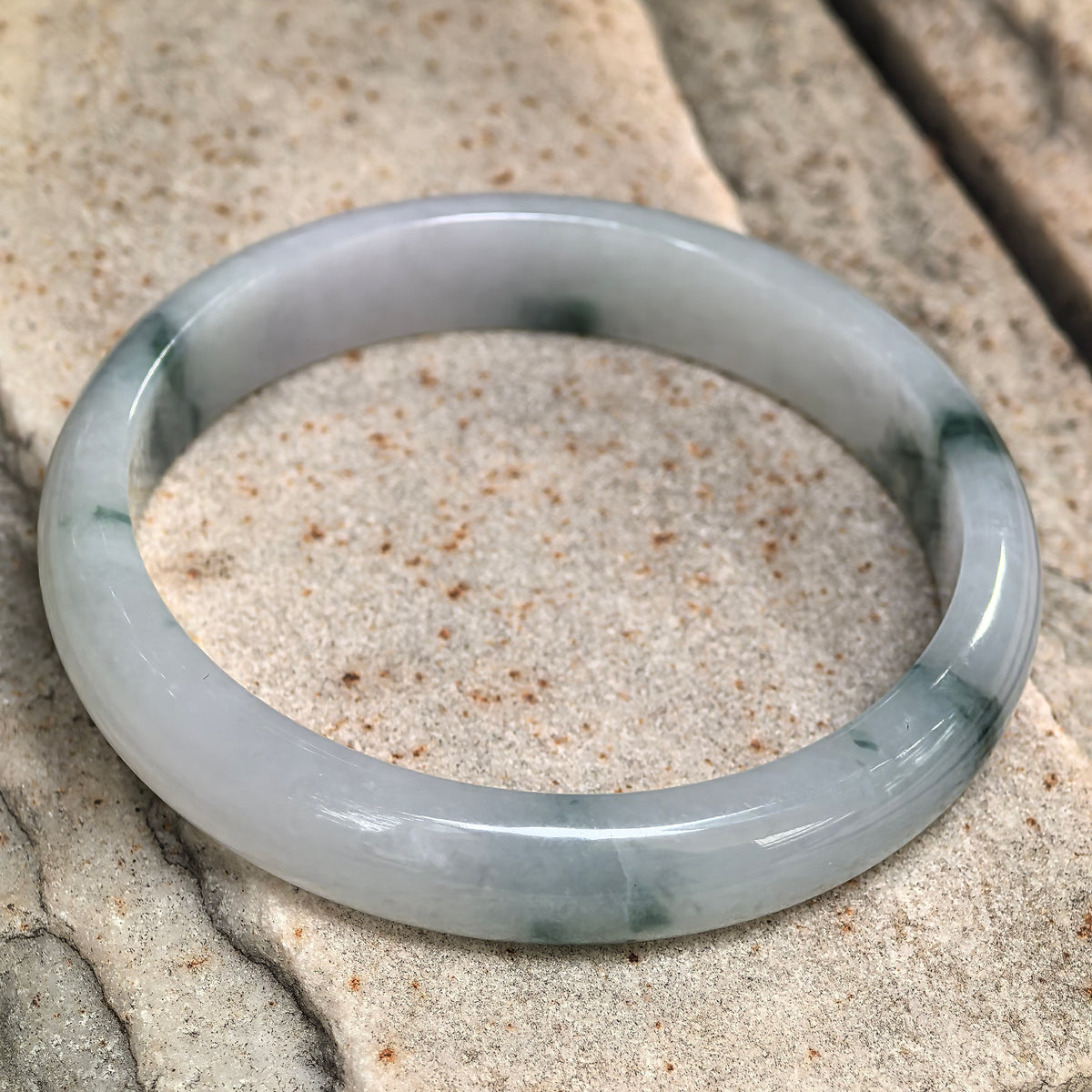 Angled view of a milky white jadeite bangle resting on stone, showing its flatter inner face and scattered green patterns.