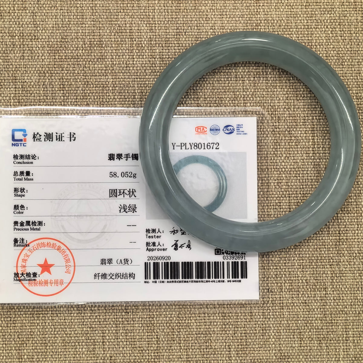 Blue-green jadeite round-bar bangle displayed with its matching NGTC certificate on a tan woven background.